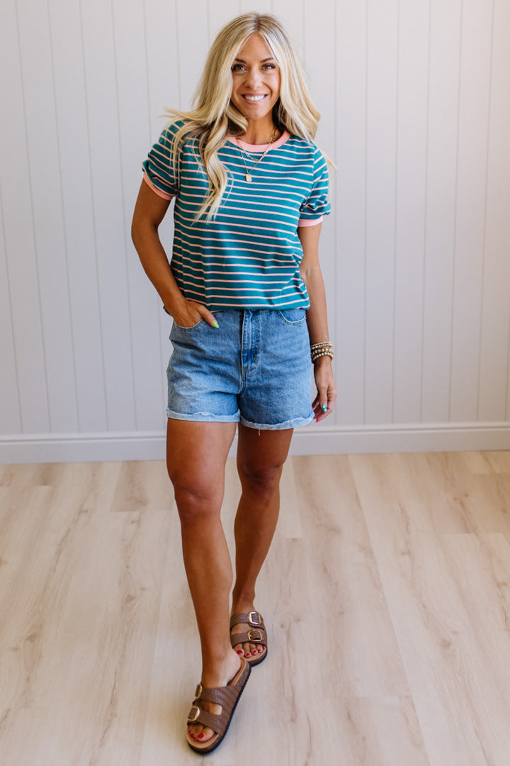 Woman wearing a striped shirt and denim shorts standing against a white paneled wall.
