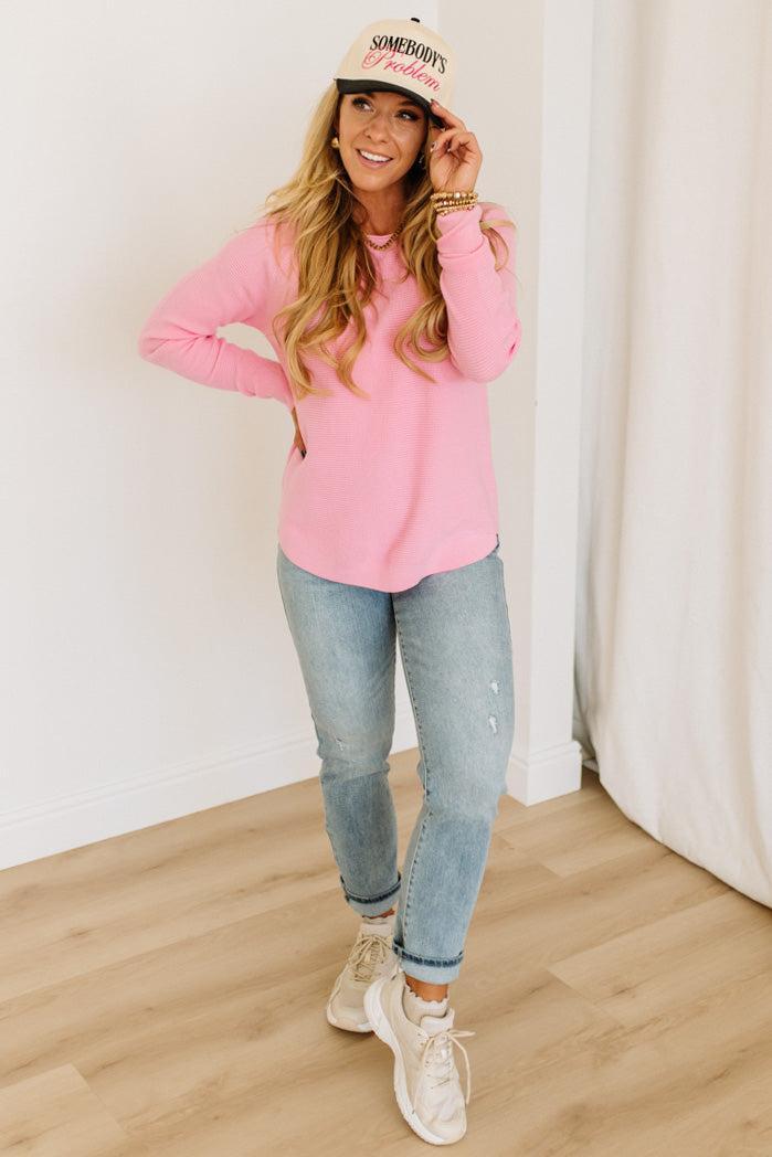 Blonde woman wearing jeans and a pink sweater with a black and white hat and sneakers.