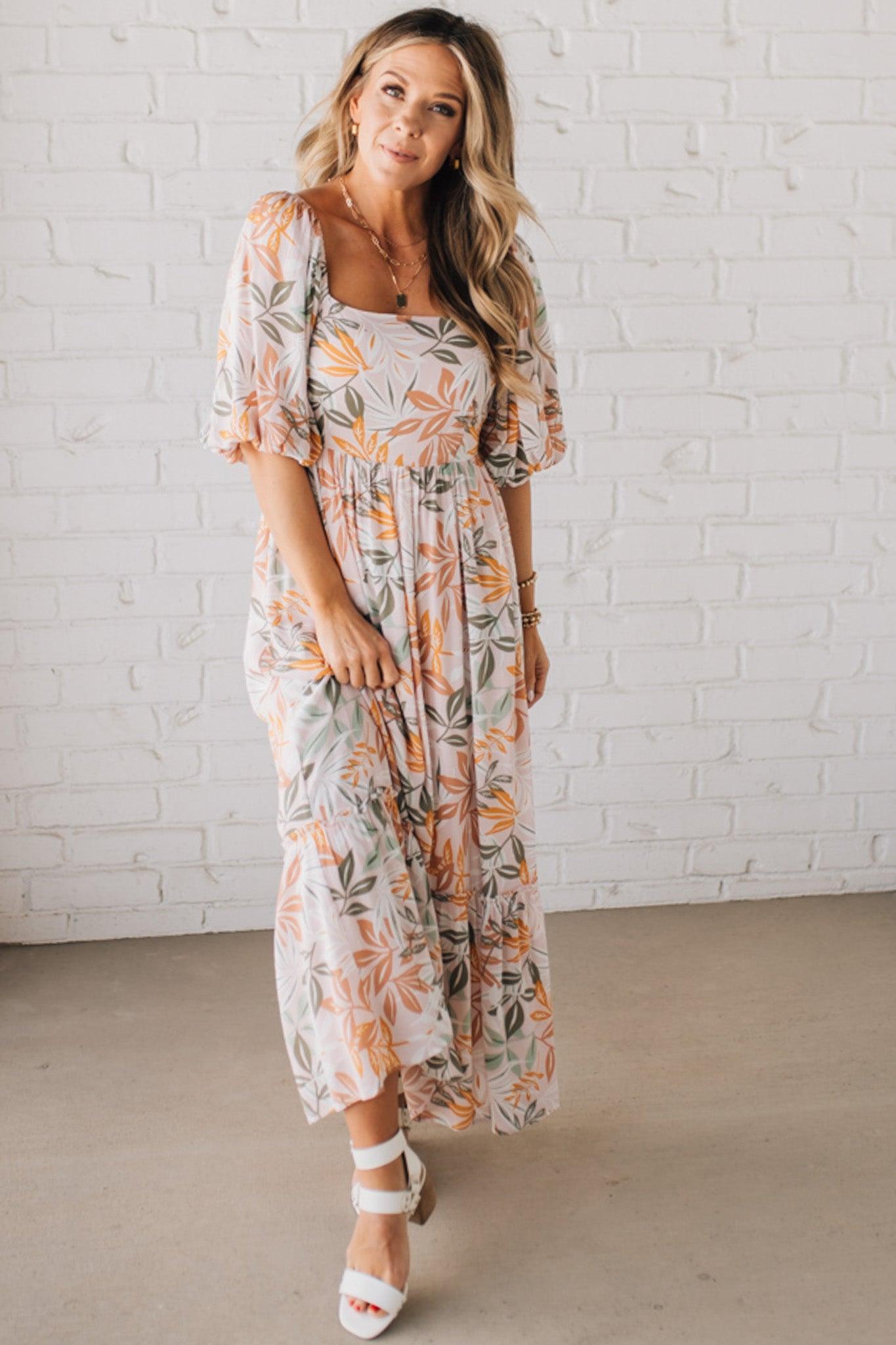 BLONDE WOMAN WEARING A MAXI DRESS WITH LEAF PRINT ON RAYON FABRIC, A SQUARE NECKLINE AND SHORT PUFF SLEEVES. HAS A SMOCKED BACK, SIDE POCKETS, AND A GATHERED HEM.