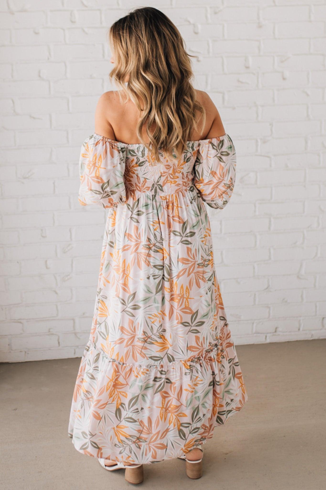 BLONDE WOMAN WEARING A MAXI DRESS WITH LEAF PRINT ON RAYON FABRIC, A SQUARE NECKLINE AND SHORT PUFF SLEEVES. HAS A SMOCKED BACK, SIDE POCKETS, AND A GATHERED HEM.