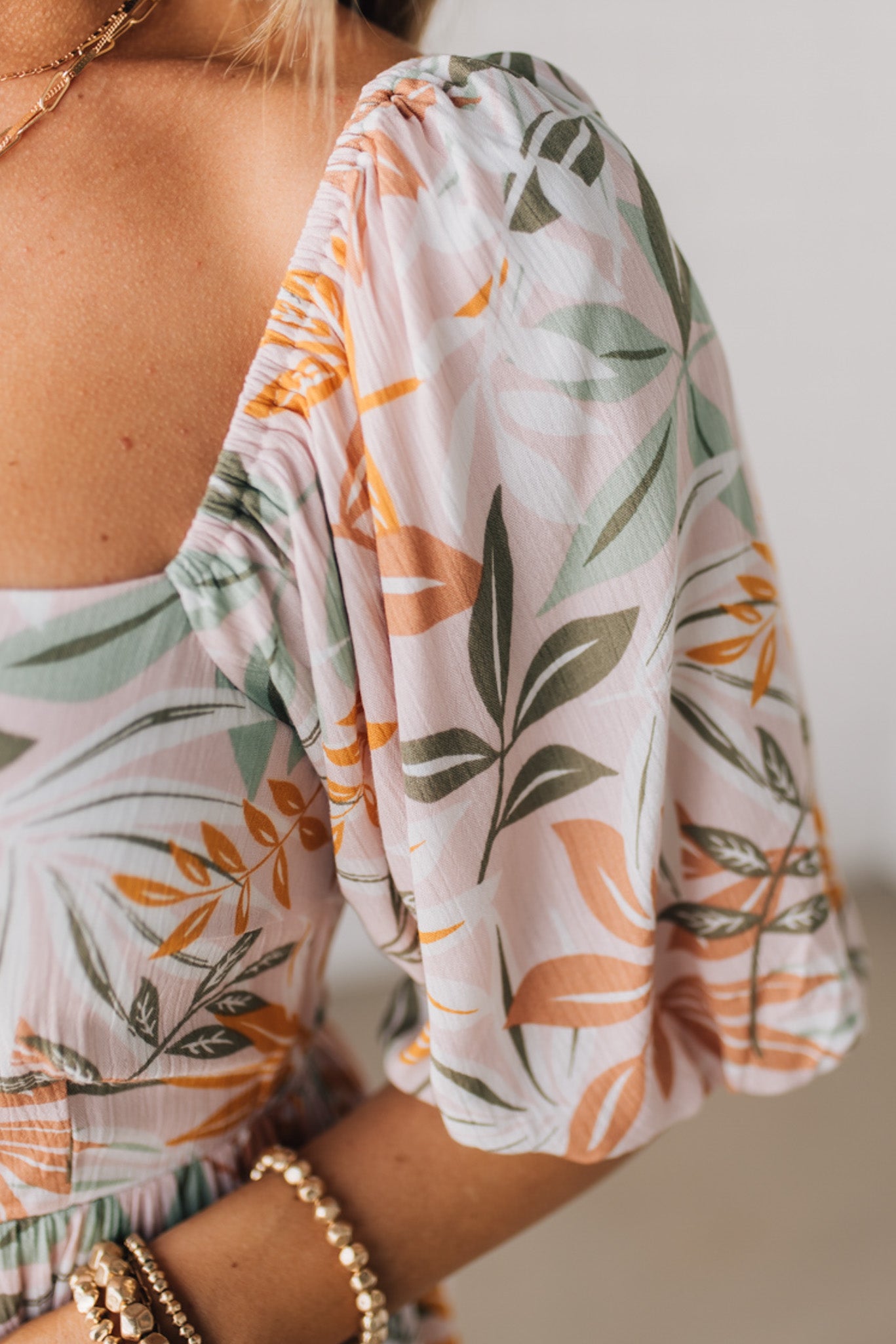 BLONDE WOMAN WEARING A MAXI DRESS WITH LEAF PRINT ON RAYON FABRIC, A SQUARE NECKLINE AND SHORT PUFF SLEEVES. HAS A SMOCKED BACK, SIDE POCKETS, AND A GATHERED HEM.