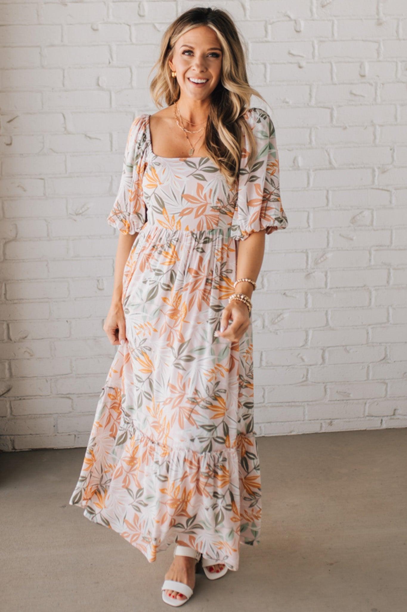 BLONDE WOMAN WEARING A MAXI DRESS WITH LEAF PRINT ON RAYON FABRIC, A SQUARE NECKLINE AND SHORT PUFF SLEEVES. HAS A SMOCKED BACK, SIDE POCKETS, AND A GATHERED HEM.