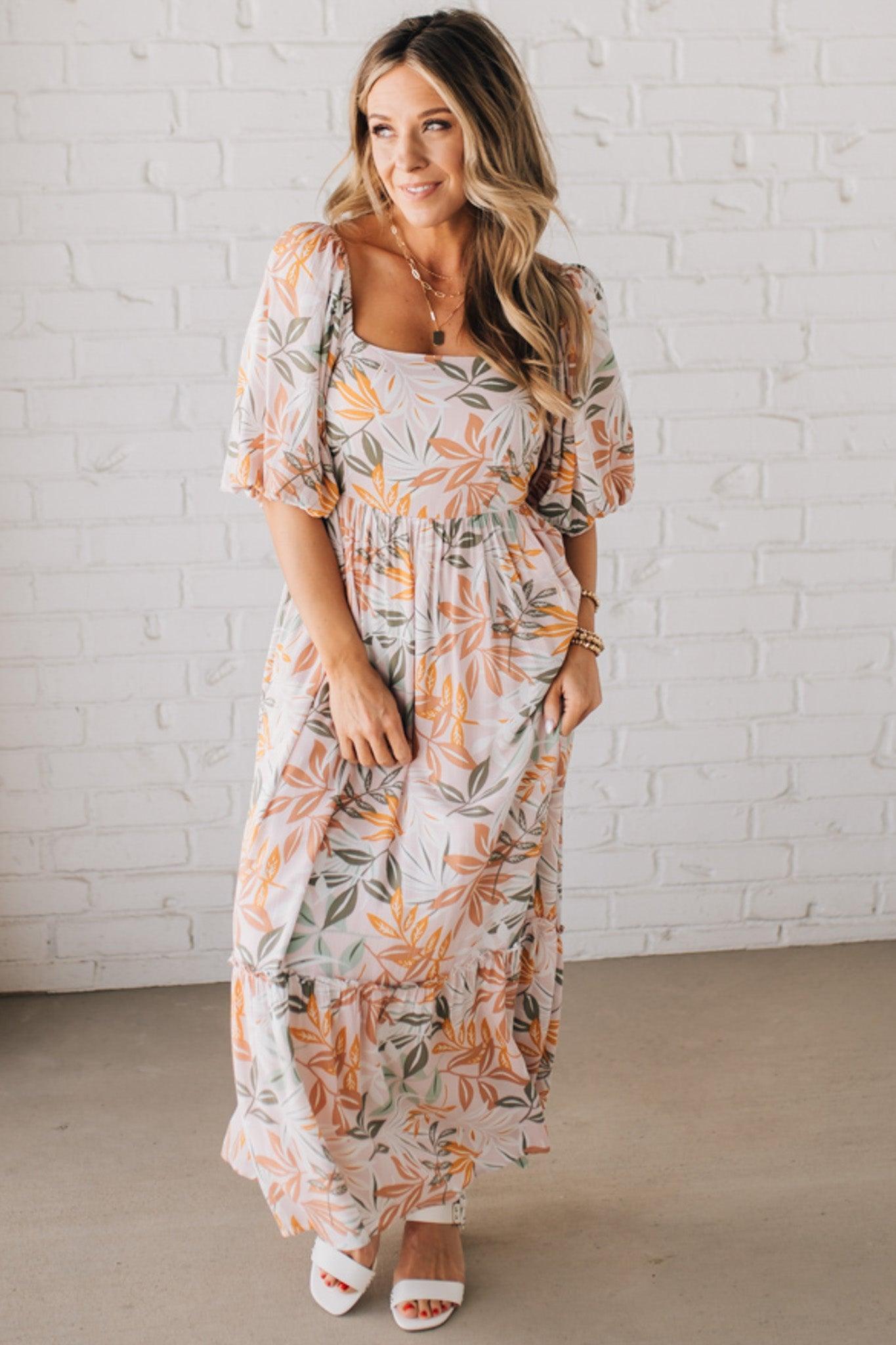 BLONDE WOMAN WEARING A MAXI DRESS WITH LEAF PRINT ON RAYON FABRIC, A SQUARE NECKLINE AND SHORT PUFF SLEEVES. HAS A SMOCKED BACK, SIDE POCKETS, AND A GATHERED HEM.