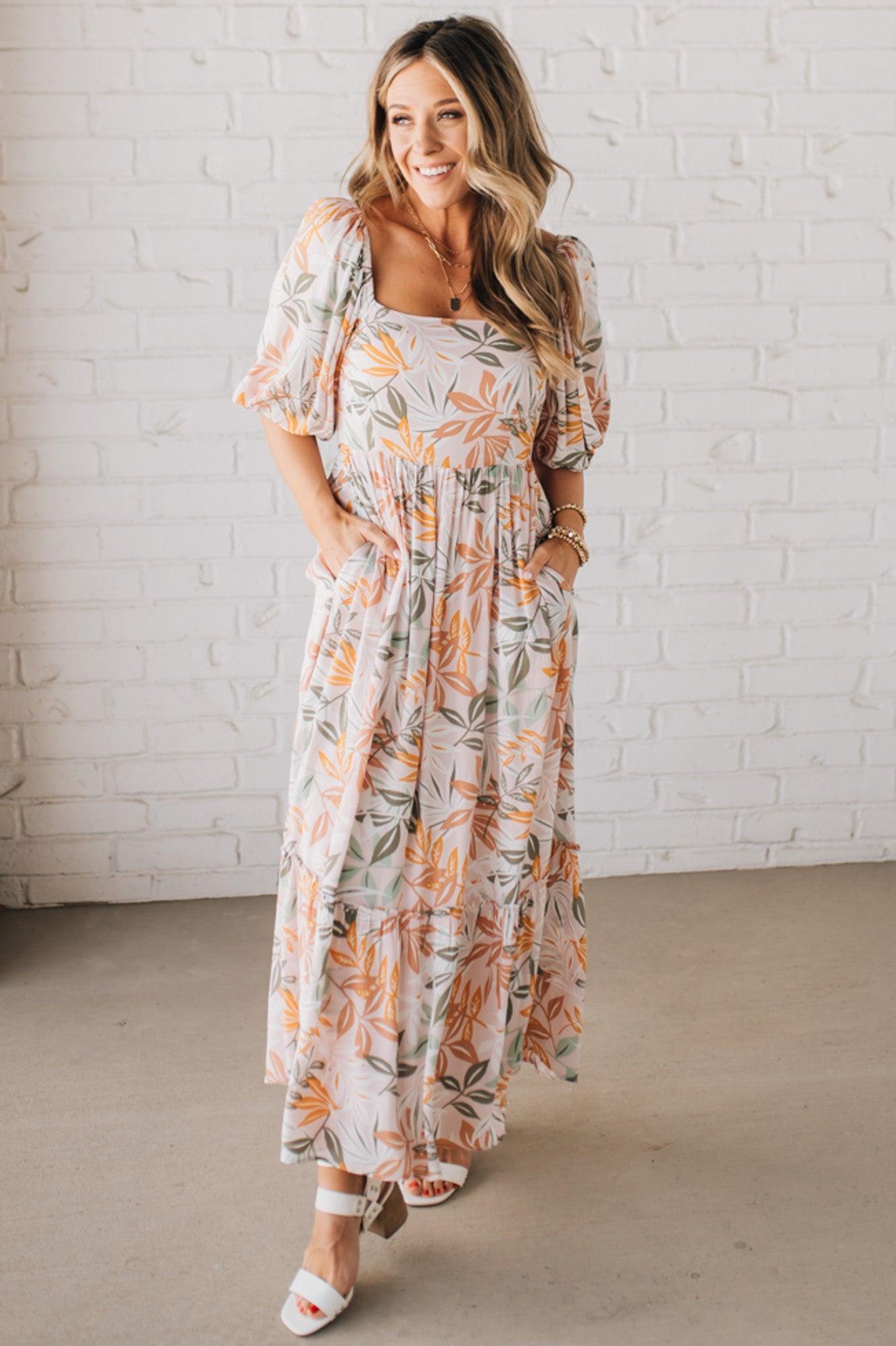 BLONDE WOMAN WEARING A MAXI DRESS WITH LEAF PRINT ON RAYON FABRIC, A SQUARE NECKLINE AND SHORT PUFF SLEEVES. HAS A SMOCKED BACK, SIDE POCKETS, AND A GATHERED HEM.