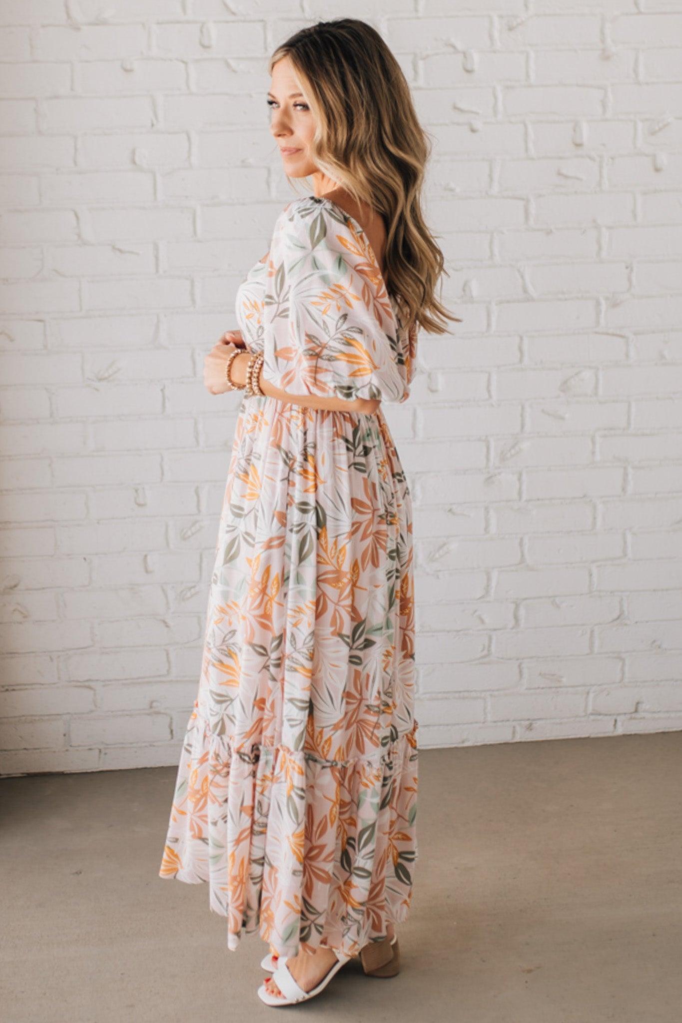BLONDE WOMAN WEARING A MAXI DRESS WITH LEAF PRINT ON RAYON FABRIC, A SQUARE NECKLINE AND SHORT PUFF SLEEVES. HAS A SMOCKED BACK, SIDE POCKETS, AND A GATHERED HEM.