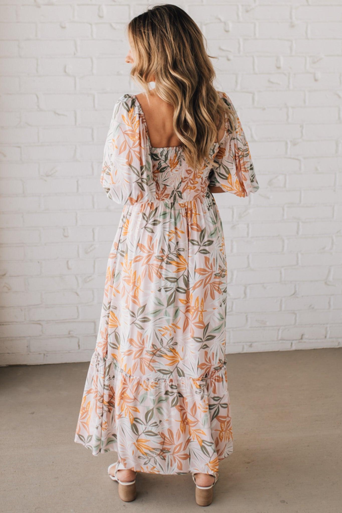 BLONDE WOMAN WEARING A MAXI DRESS WITH LEAF PRINT ON RAYON FABRIC, A SQUARE NECKLINE AND SHORT PUFF SLEEVES. HAS A SMOCKED BACK, SIDE POCKETS, AND A GATHERED HEM.