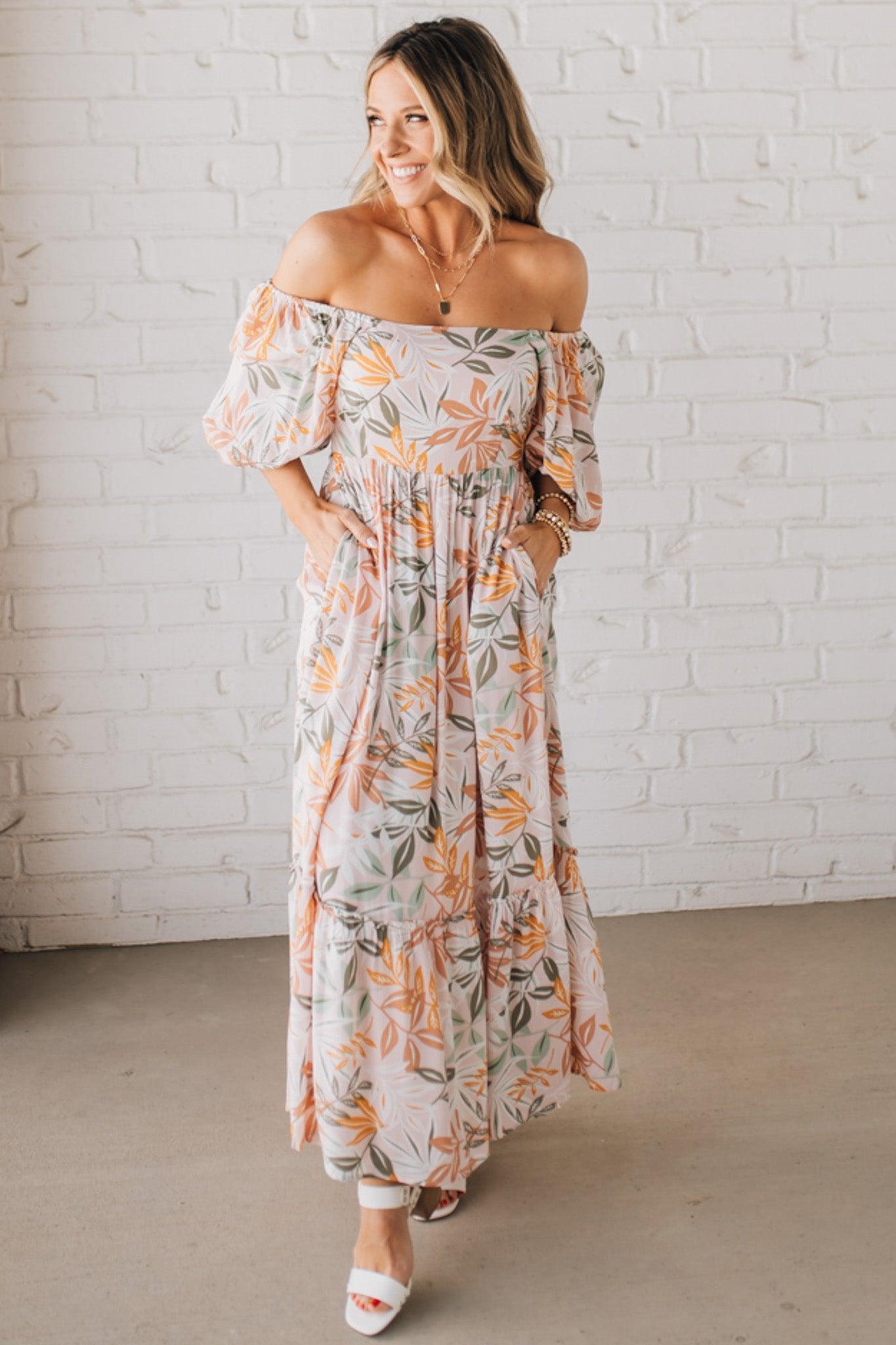 BLONDE WOMAN WEARING A MAXI DRESS WITH LEAF PRINT ON RAYON FABRIC, A SQUARE NECKLINE AND SHORT PUFF SLEEVES. HAS A SMOCKED BACK, SIDE POCKETS, AND A GATHERED HEM.