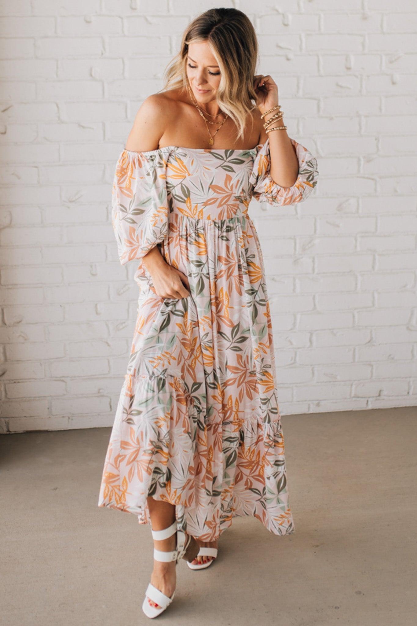 BLONDE WOMAN WEARING A MAXI DRESS WITH LEAF PRINT ON RAYON FABRIC, A SQUARE NECKLINE AND SHORT PUFF SLEEVES. HAS A SMOCKED BACK, SIDE POCKETS, AND A GATHERED HEM.
