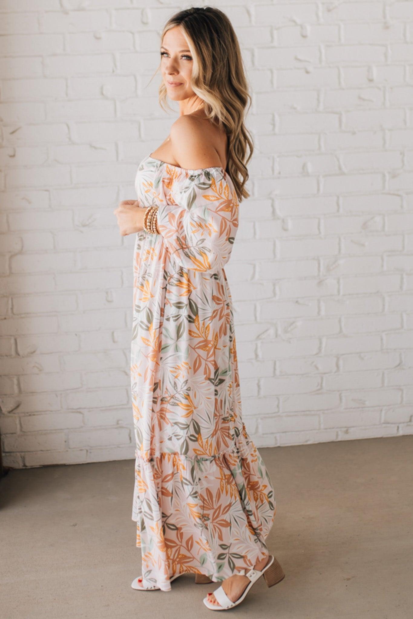 BLONDE WOMAN WEARING A MAXI DRESS WITH LEAF PRINT ON RAYON FABRIC, A SQUARE NECKLINE AND SHORT PUFF SLEEVES. HAS A SMOCKED BACK, SIDE POCKETS, AND A GATHERED HEM.