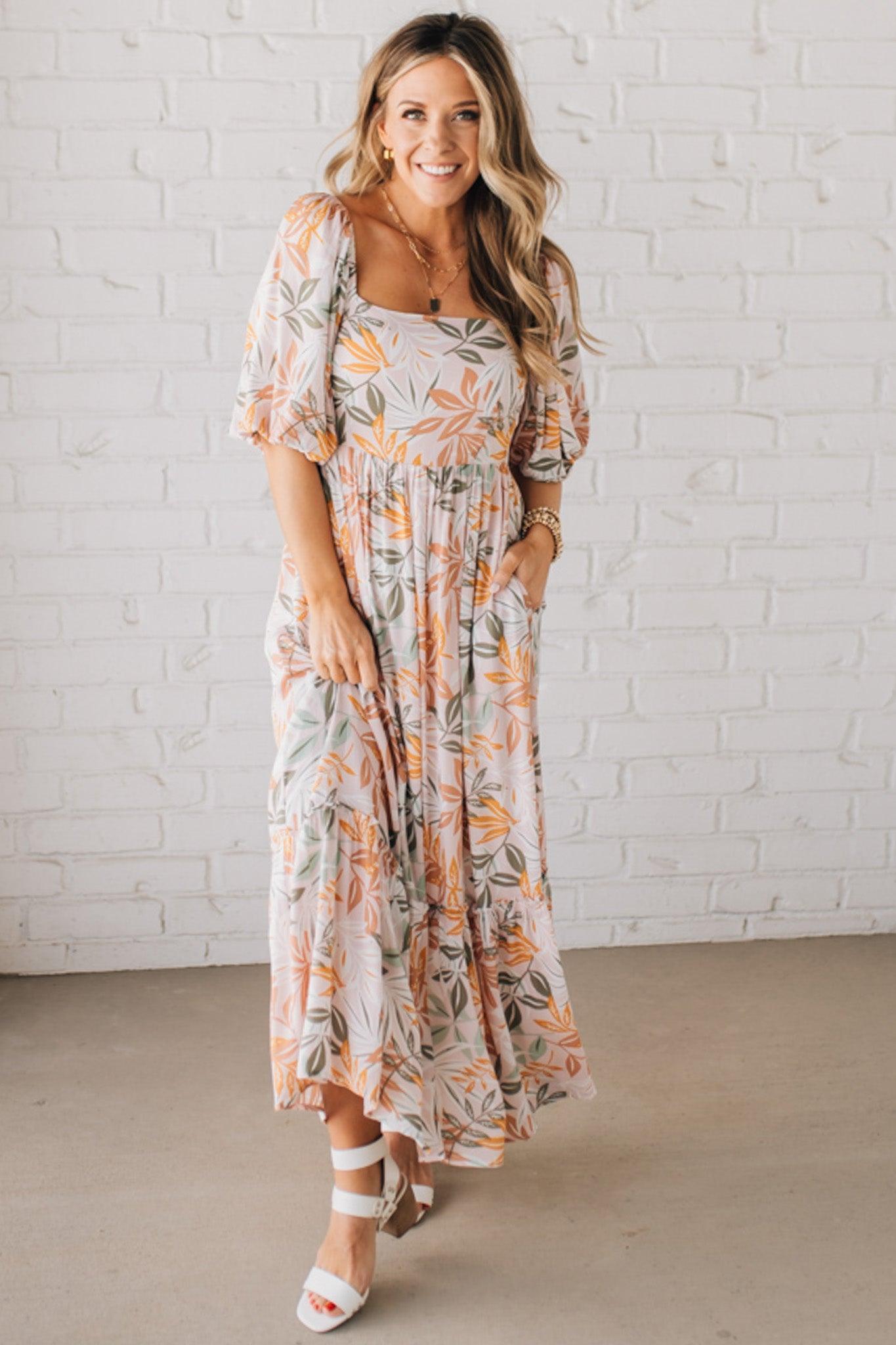 BLONDE WOMAN WEARING A MAXI DRESS WITH LEAF PRINT ON RAYON FABRIC, A SQUARE NECKLINE AND SHORT PUFF SLEEVES. HAS A SMOCKED BACK, SIDE POCKETS, AND A GATHERED HEM.