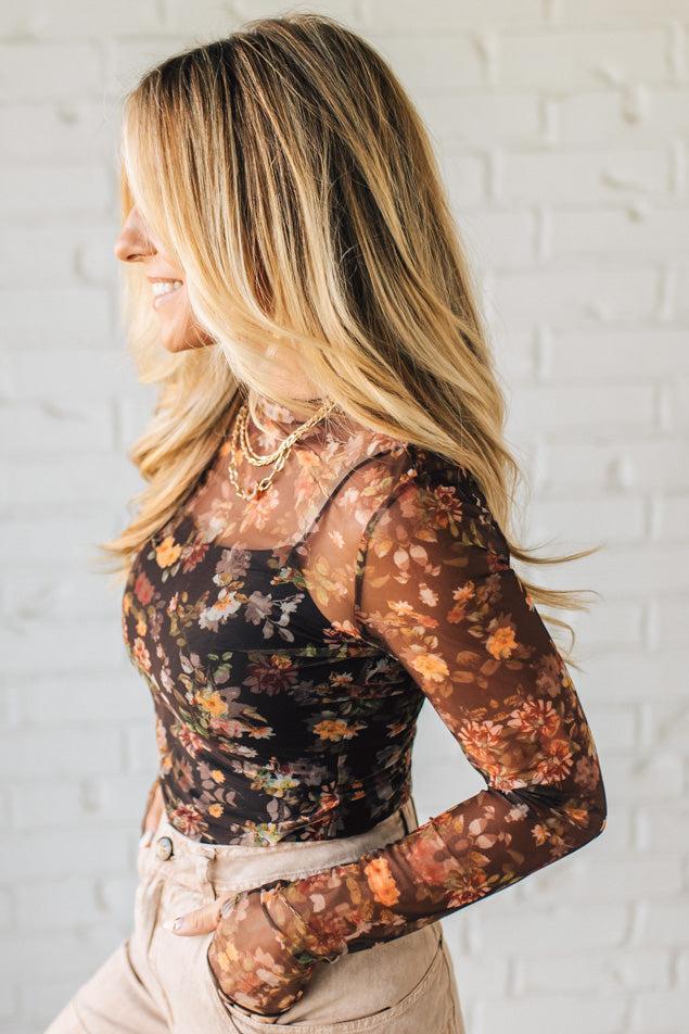 Woman wearing a floral long-sleeve top against a white brick wall.