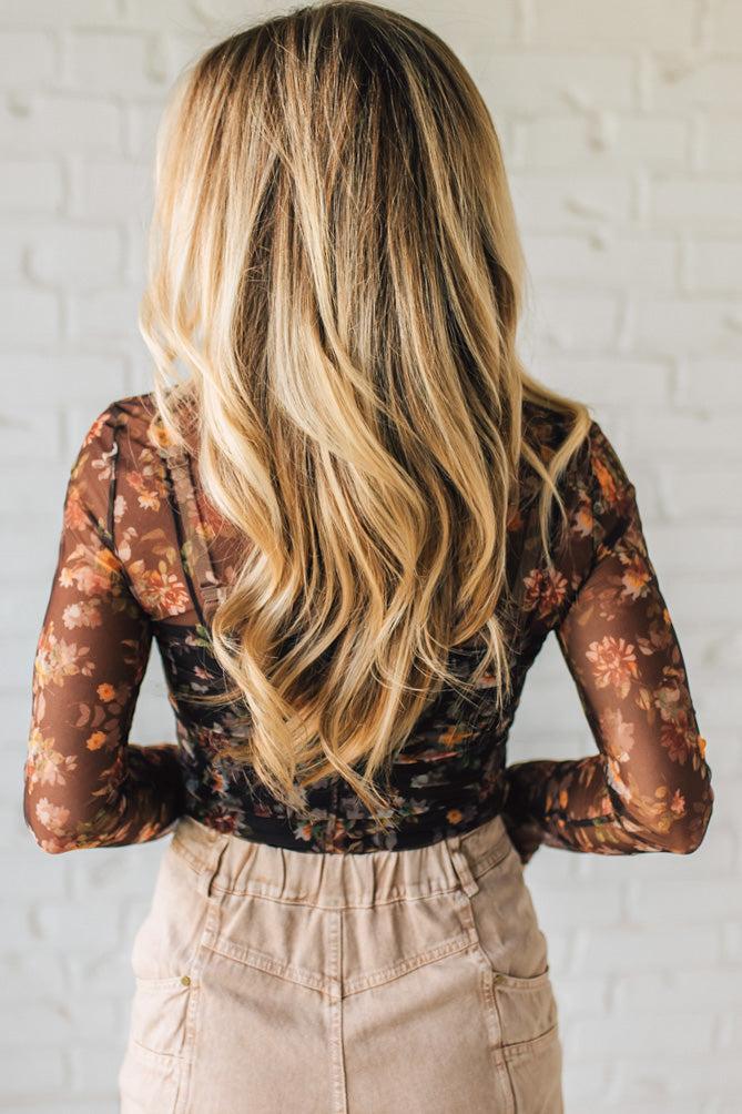 Person wearing a floral long-sleeve top and beige pants against a white brick wall.