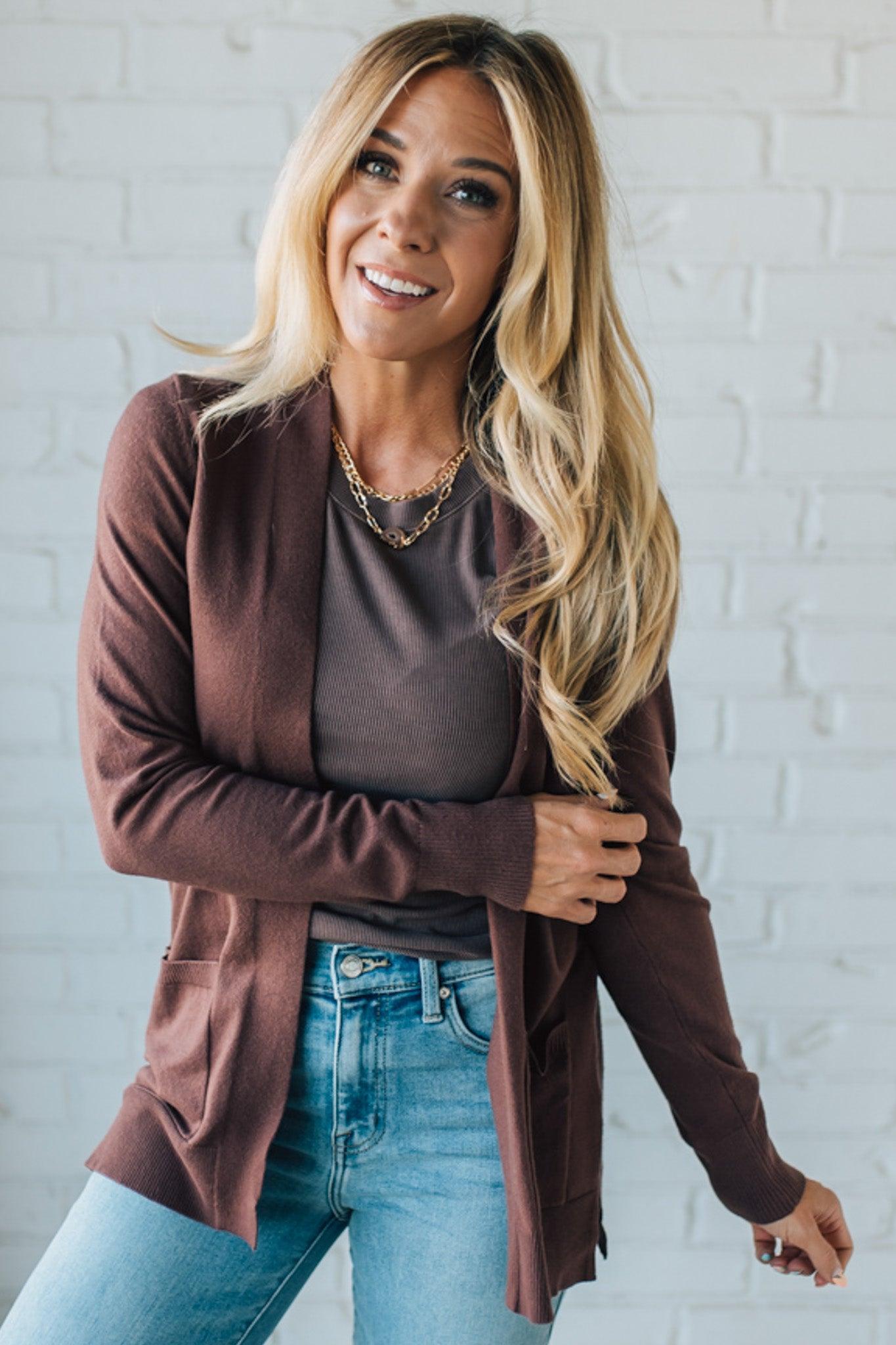 Woman wearing a brown cardigan over a gray shirt with blue jeans against a white brick wall.