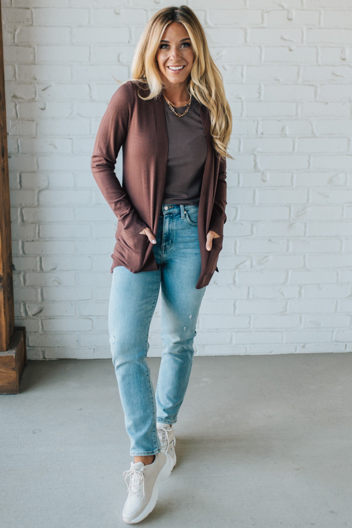 Woman wearing a brown cardigan, brown top, light blue jeans, and white sneakers against a white brick wall.