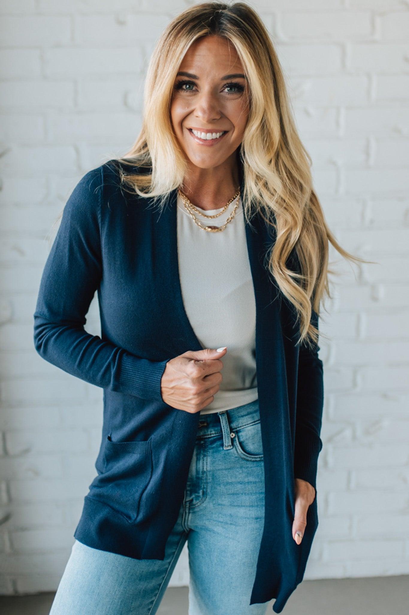Woman wearing a navy cardigan, white top, and blue jeans against a white brick wall.