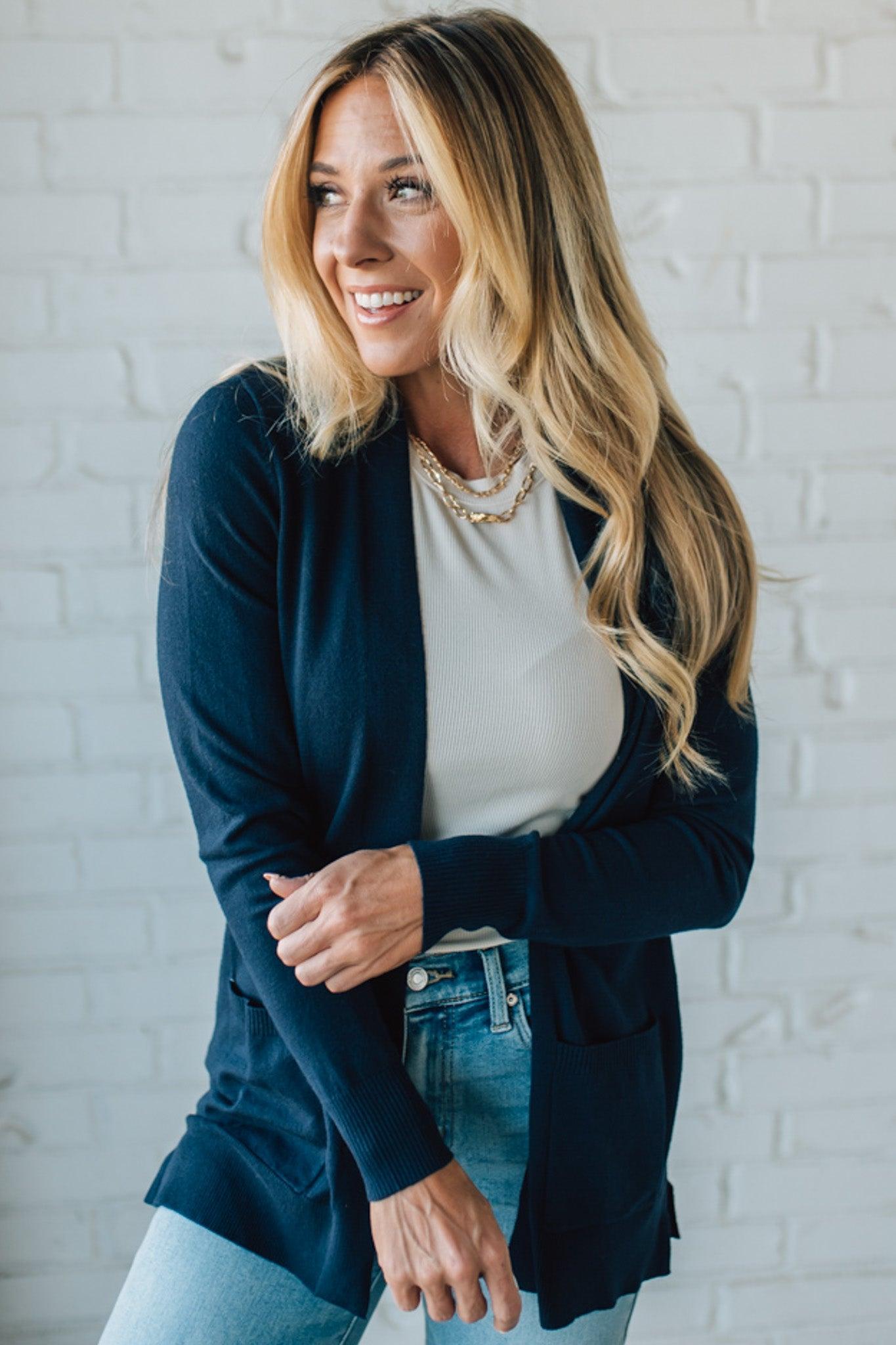 Woman wearing a navy cardigan over a white top and blue jeans against a white brick wall.