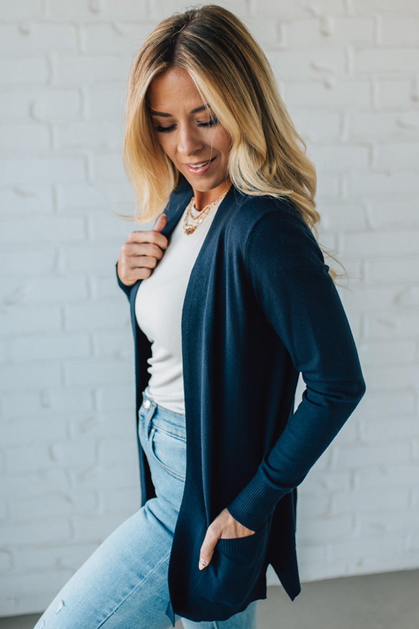 Woman wearing a navy blazer over a white top and light blue jeans against a white brick wall.
