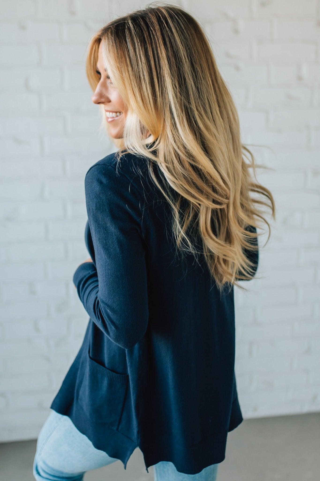 Woman wearing a dark blue cardigan against a light gray brick wall.