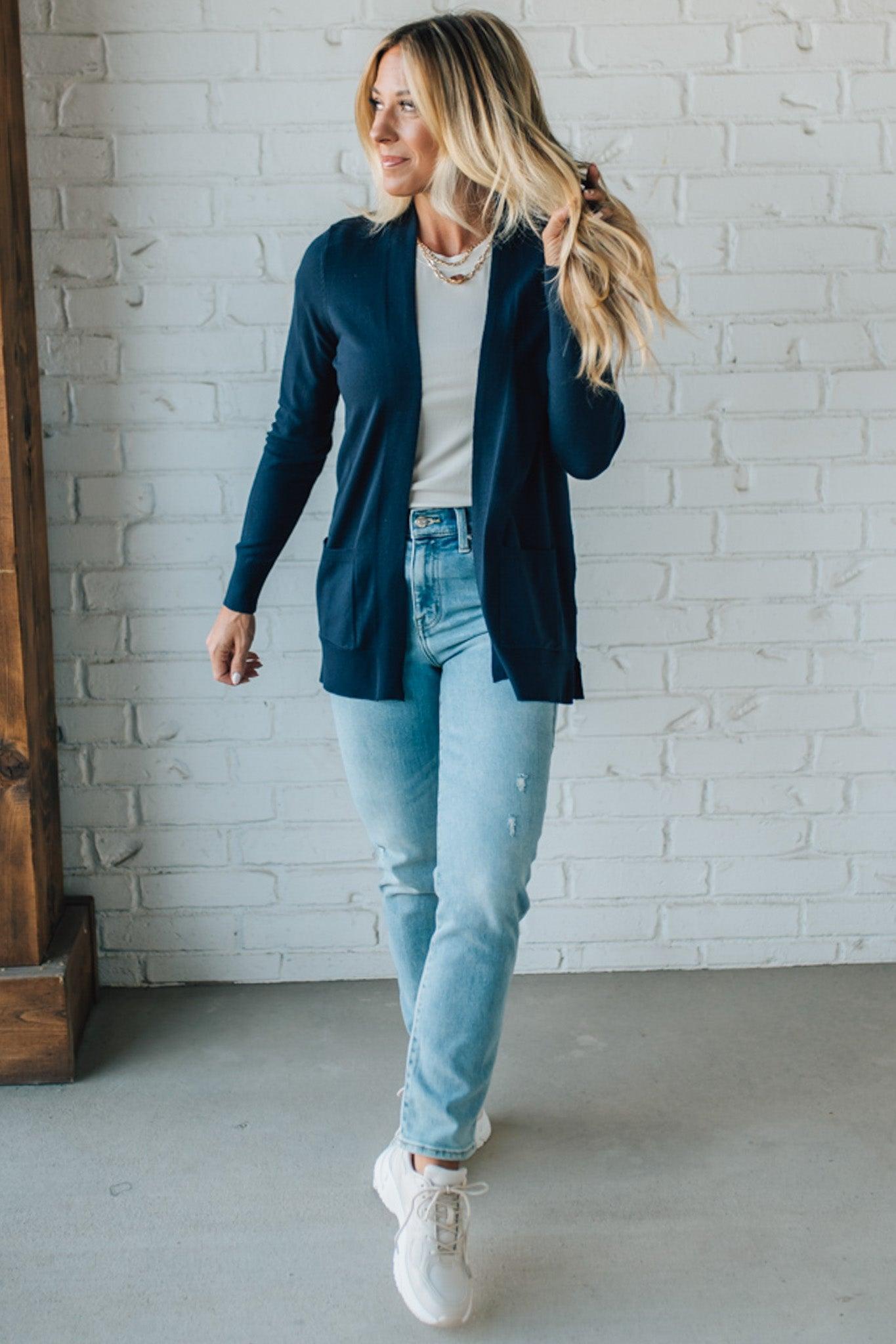Woman wearing a navy cardigan, white shirt, light blue jeans, and white sneakers against a white brick wall.