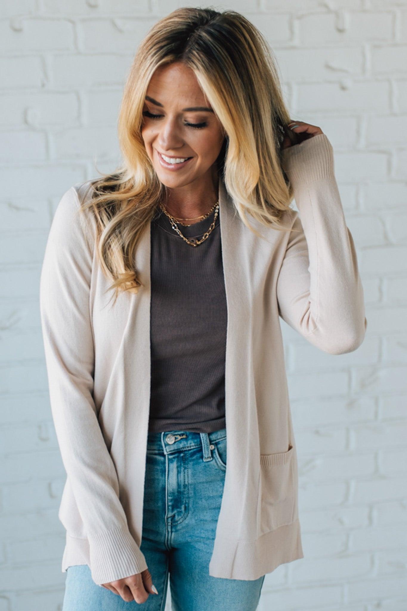 Woman wearing a beige cardigan over a gray shirt and blue jeans against a white brick wall.