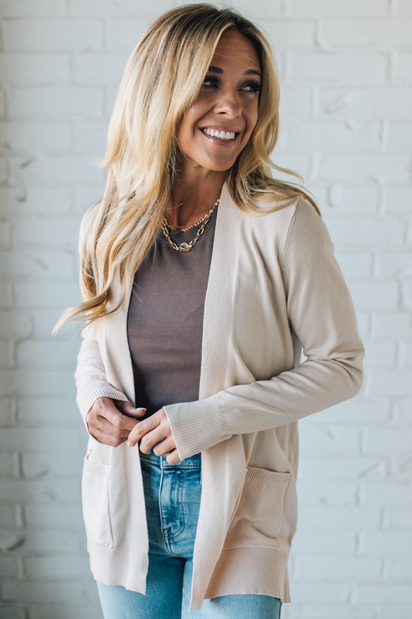 Woman wearing a beige cardigan over a gray top and blue jeans against a white brick wall.