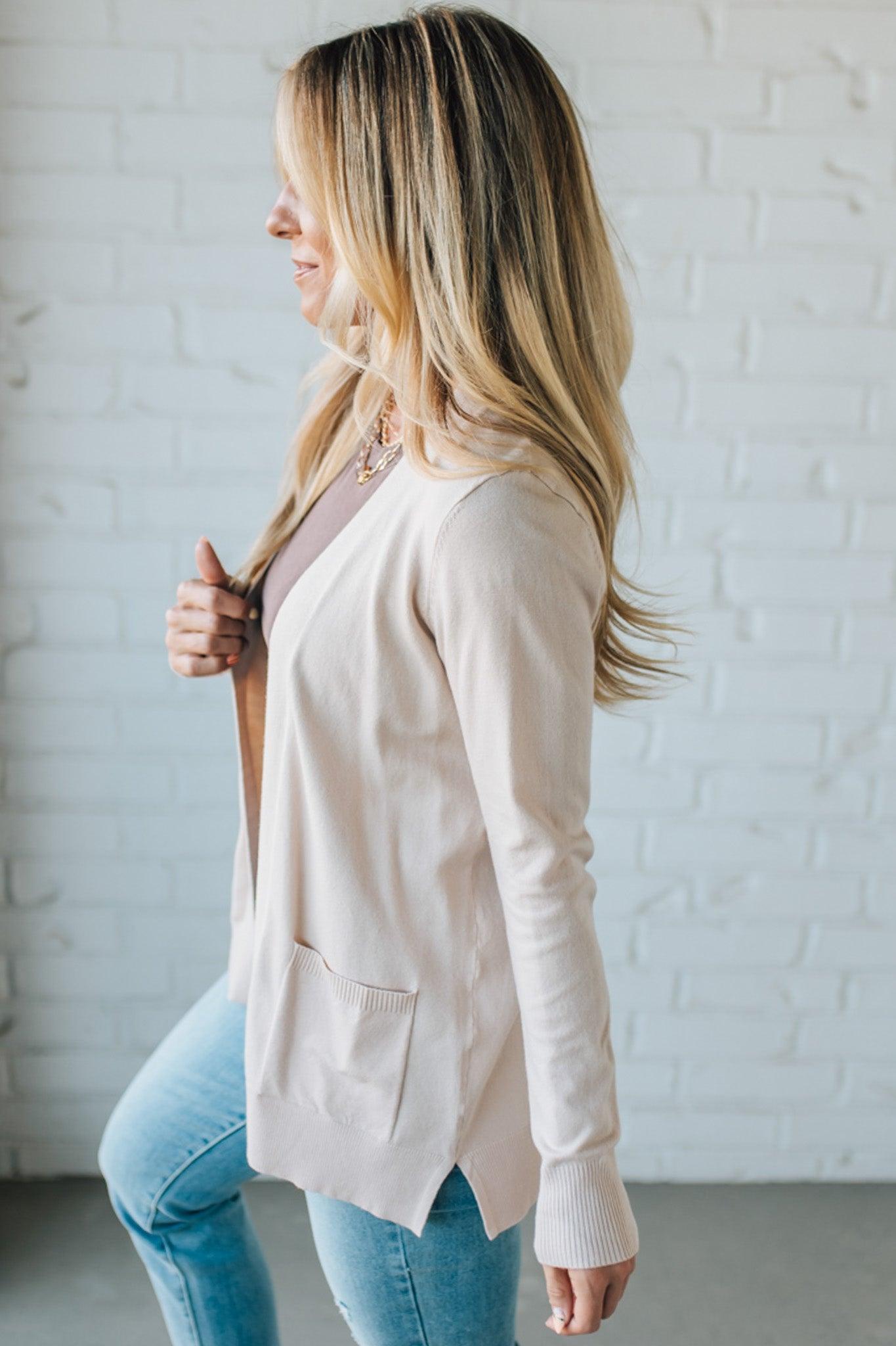 Woman wearing a beige cardigan and blue jeans against a white brick wall.