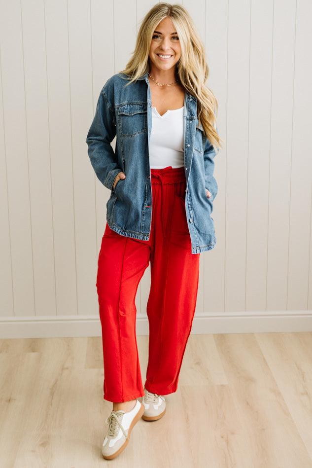 Woman wearing a denim jacket, white top, and red pants standing against a light wooden paneled wall.