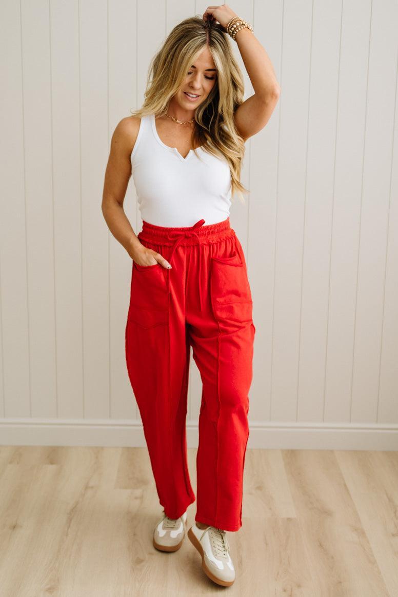 Woman wearing a white top and red pants standing against a light wooden paneled wall.