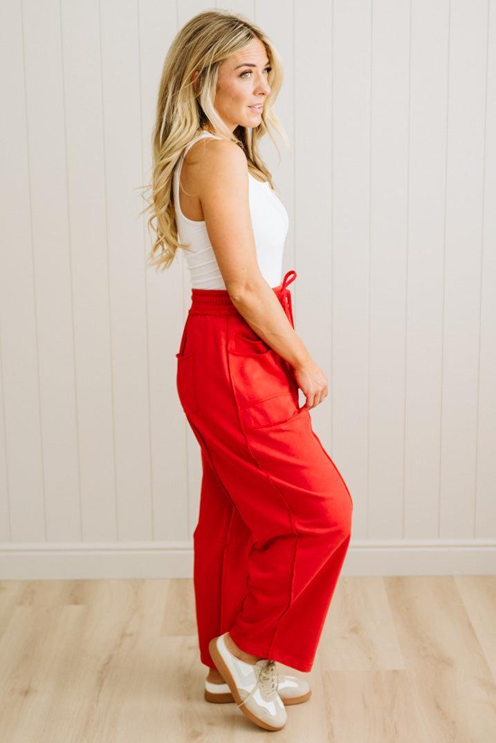 Woman wearing a white top and red pants standing against a light wooden paneled wall.