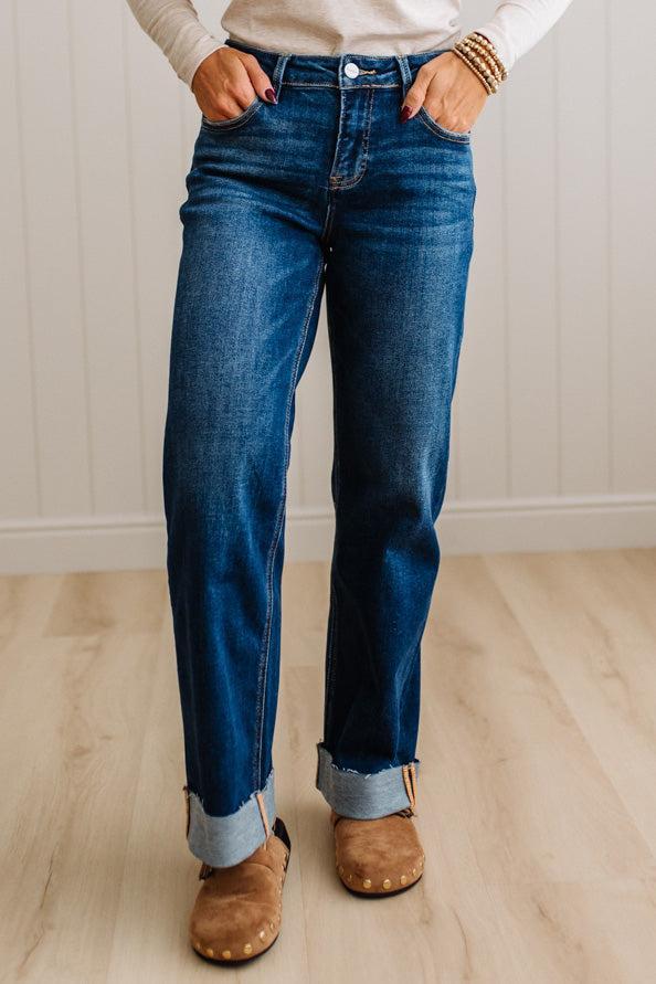 A person wearing dark blue straight-leg denim jeans with rolled cuffs, paired with a cream long-sleeve top and brown suede mule shoes, standing on a light wood floor against a white paneled wall.