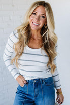 Woman wearing a striped long-sleeve top and blue jeans against a white brick wall.