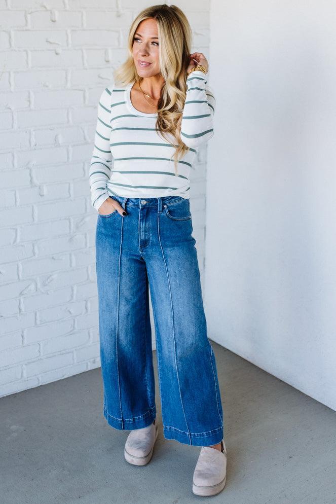 Woman wearing a striped sweater and wide-leg jeans against a white brick wall.