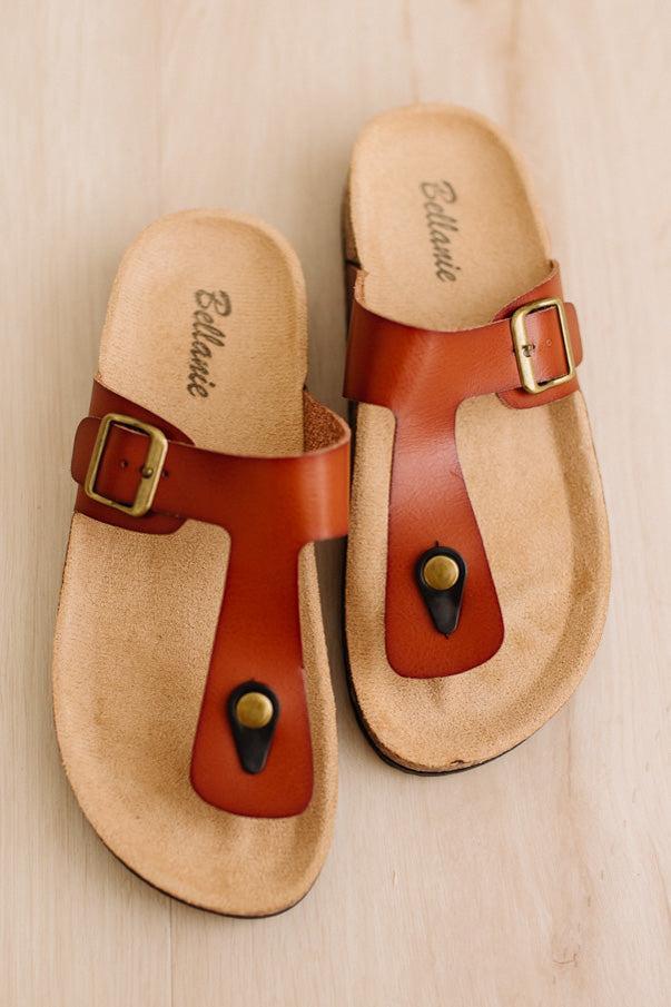A pair of brown leather thong sandals with gold-tone buckles and metal eyelets, laid flat on a light wood background. The brand name 'Ellamae' is printed on the footbeds.