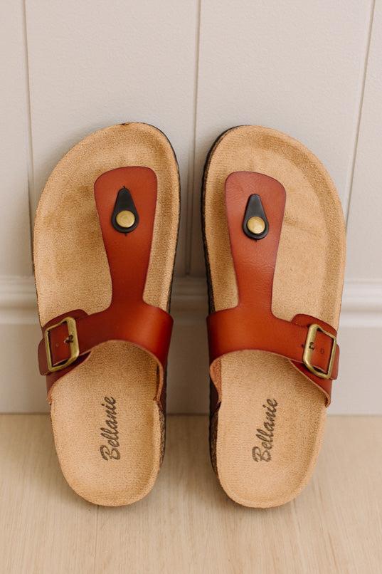 A pair of Birkenstock Bellemanie slide sandals with tan cork footbeds and reddish-brown leather straps, displayed on a light wood floor against a white paneled wall.