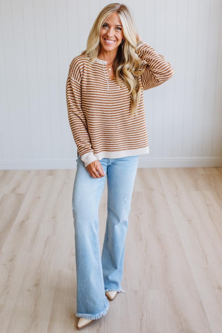 A woman modeling a brown and cream striped long-sleeve henley sweater with light-wash blue flare jeans, standing in a room with light wood floors and white paneled walls.
