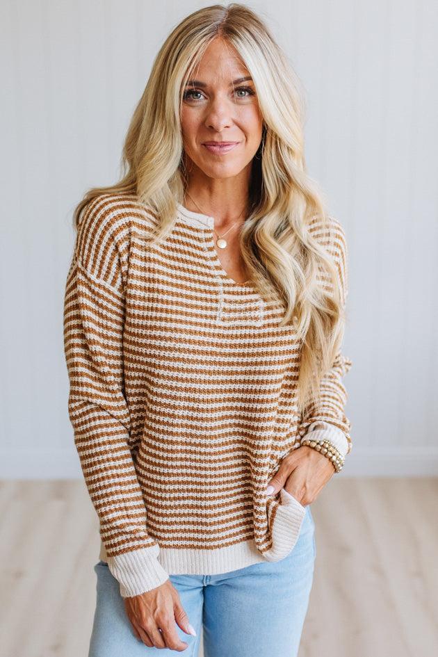 A blonde woman wearing a brown and cream horizontally striped waffle-knit sweater with a henley neckline, paired with light blue jeans, standing against a white paneled wall.