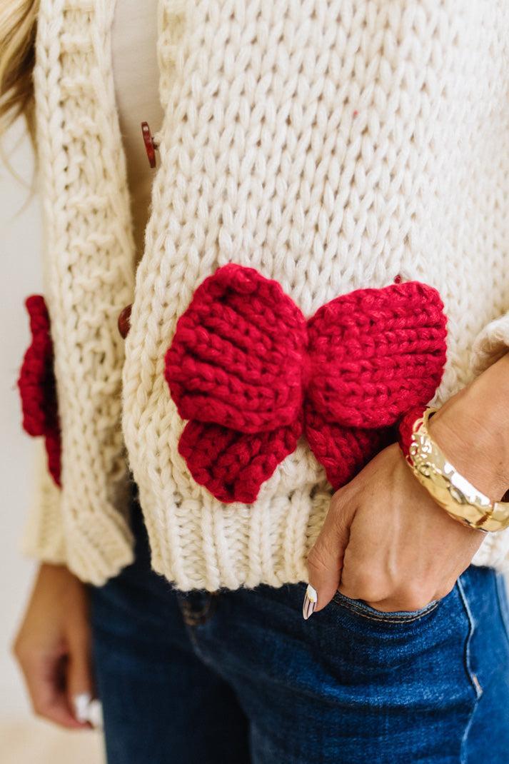 Blonde Woman in a Bright Studio wearing a cream Front Button Closure chunky sweater with red Bow Trim, Long Sleeves.