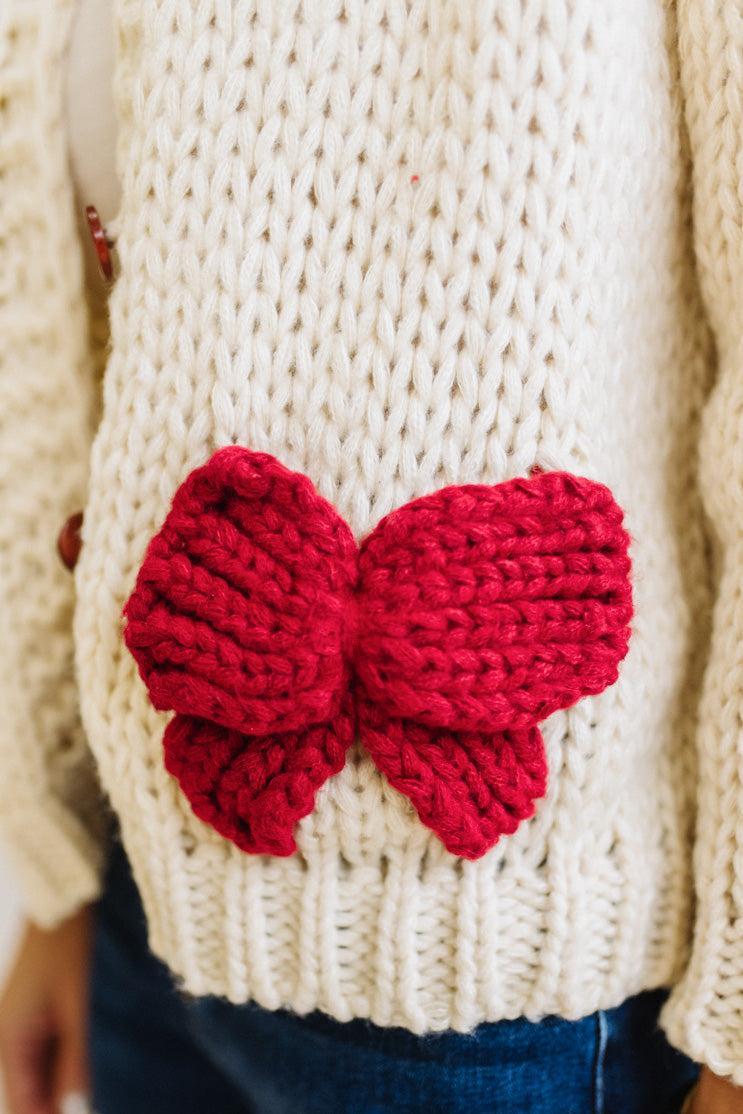 Blonde Woman in a Bright Studio wearing a cream Front Button Closure chunky sweater with red Bow Trim, Long Sleeves.