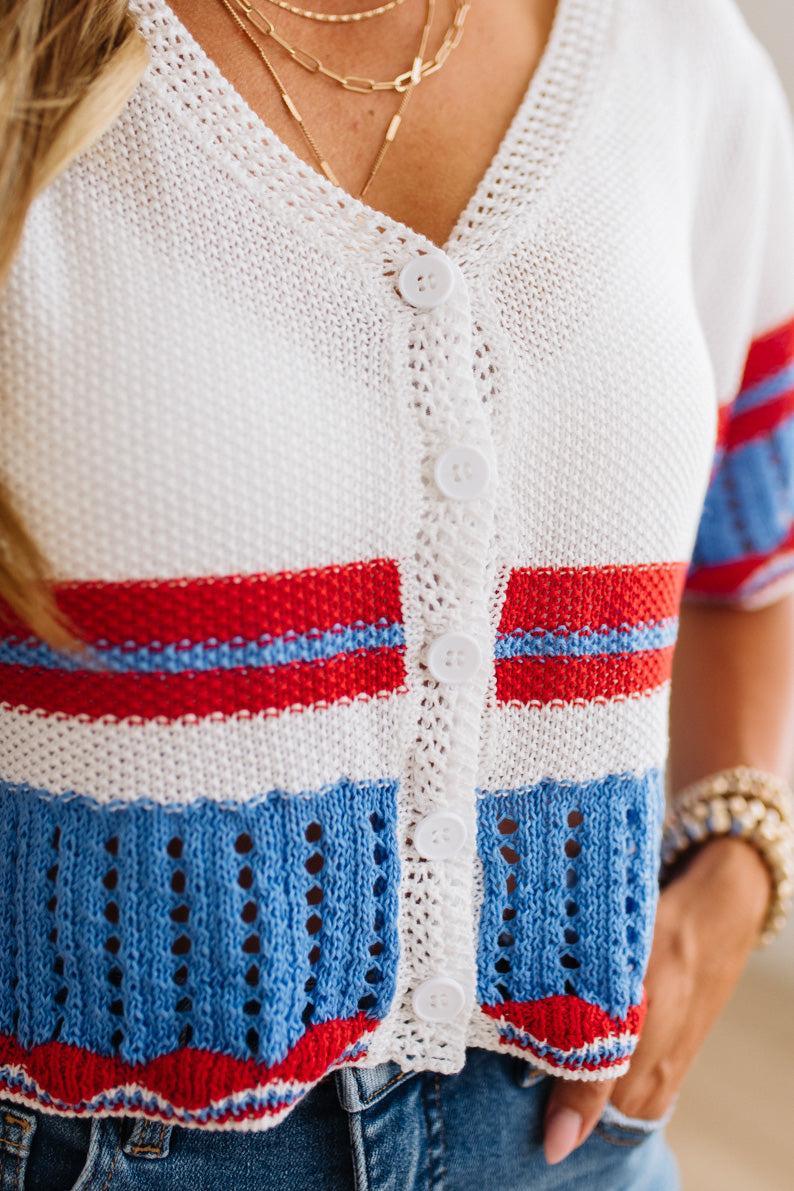 A close-up of a woman wearing a white knit cardigan with red and blue horizontal stripes, paired with blue jeans and layered gold necklaces.