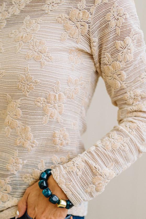 Blonde Woman Wearing A Floral Embroidered Stretch Lace Long Sleeve Top with Dark Jeans and Light Brown Belt.