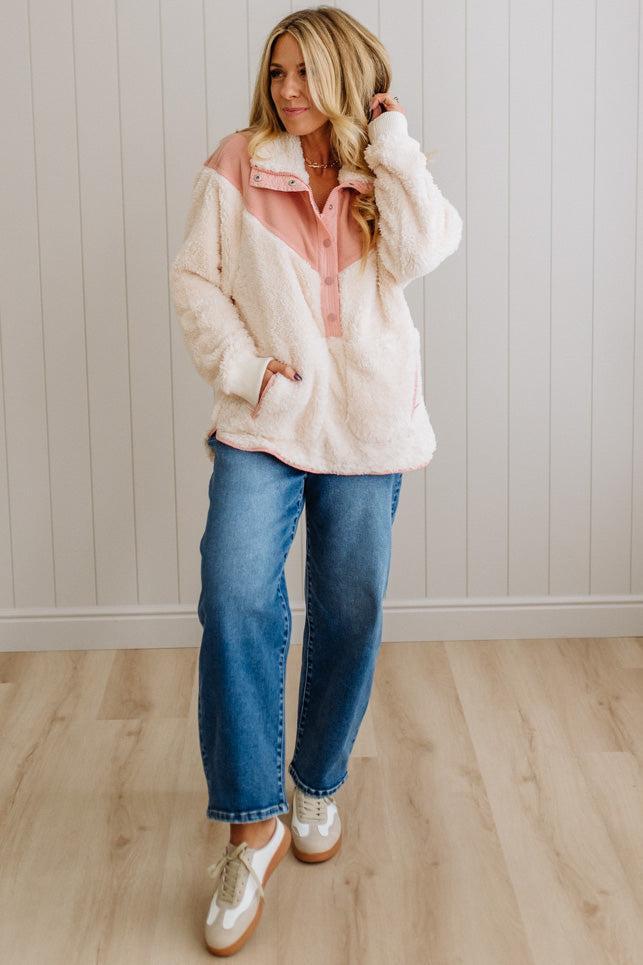 Woman wearing a white fleece jacket with pink interior, blue jeans, and white sneakers in a room with wooden flooring and a light-colored wall.