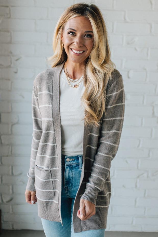 Woman wearing a gray striped cardigan over a white shirt and blue jeans, standing against a white brick wall.