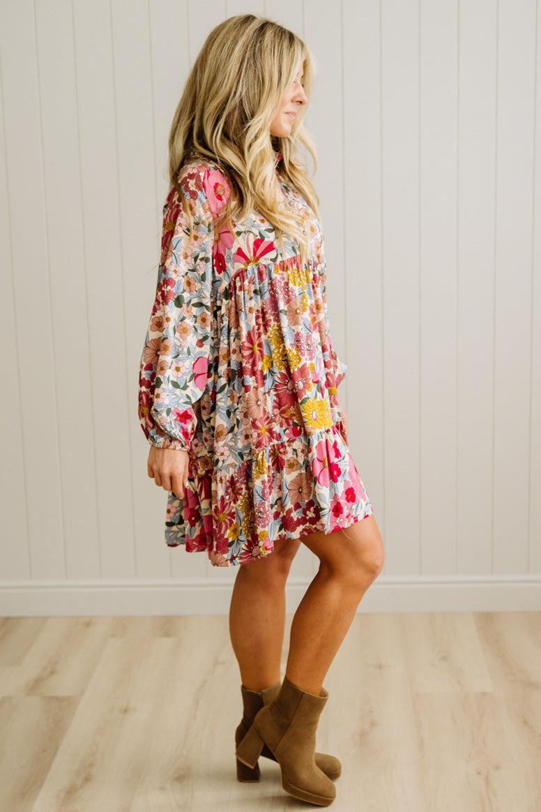 Woman wearing a colorful floral dress with long sleeves against a white paneled wall.