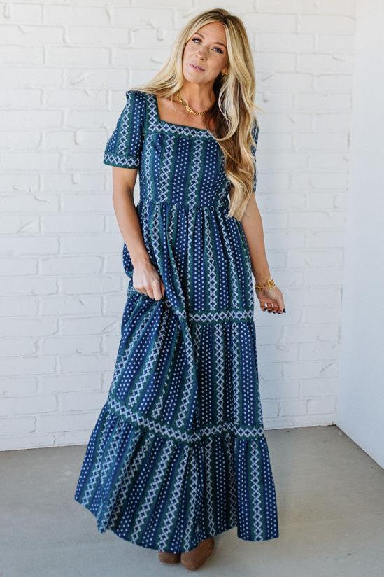 Woman wearing a blue patterned dress against a white brick wall.