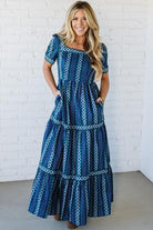 Woman wearing a blue patterned dress against a white brick wall.