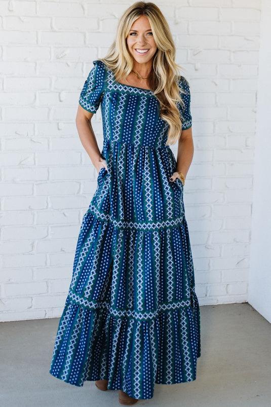 Woman wearing a blue patterned dress against a white brick wall.