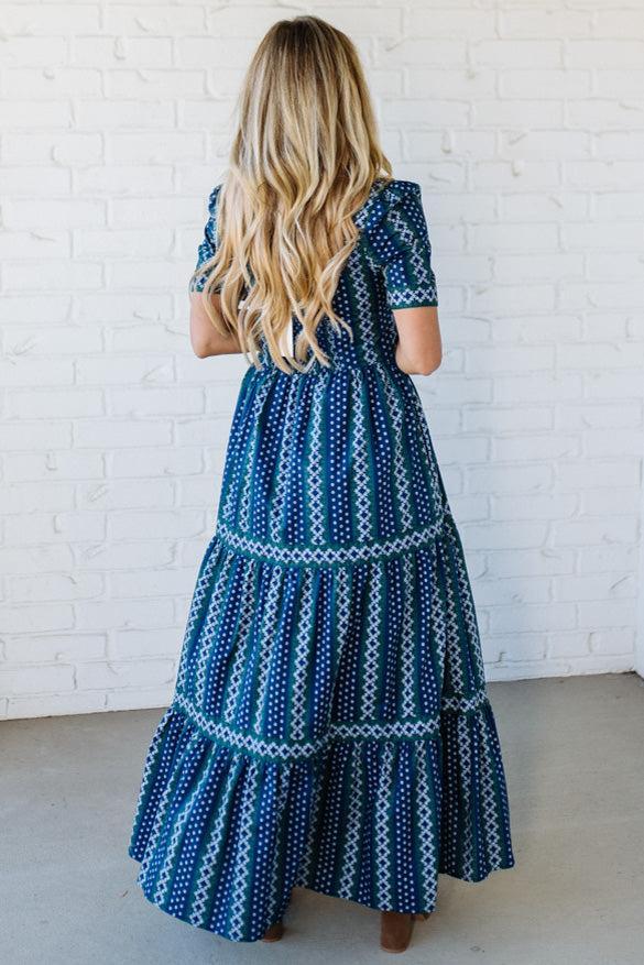 Woman wearing a blue patterned dress against a white brick wall.