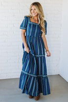 Woman wearing a blue patterned dress against a white brick wall.