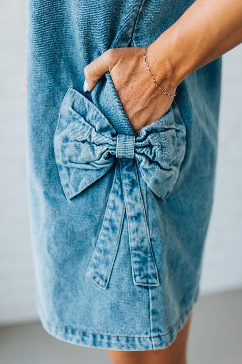Blonde woman wearing a denim shift dress to the knee with short sleeves, side pickets and bow accents on each pocket.