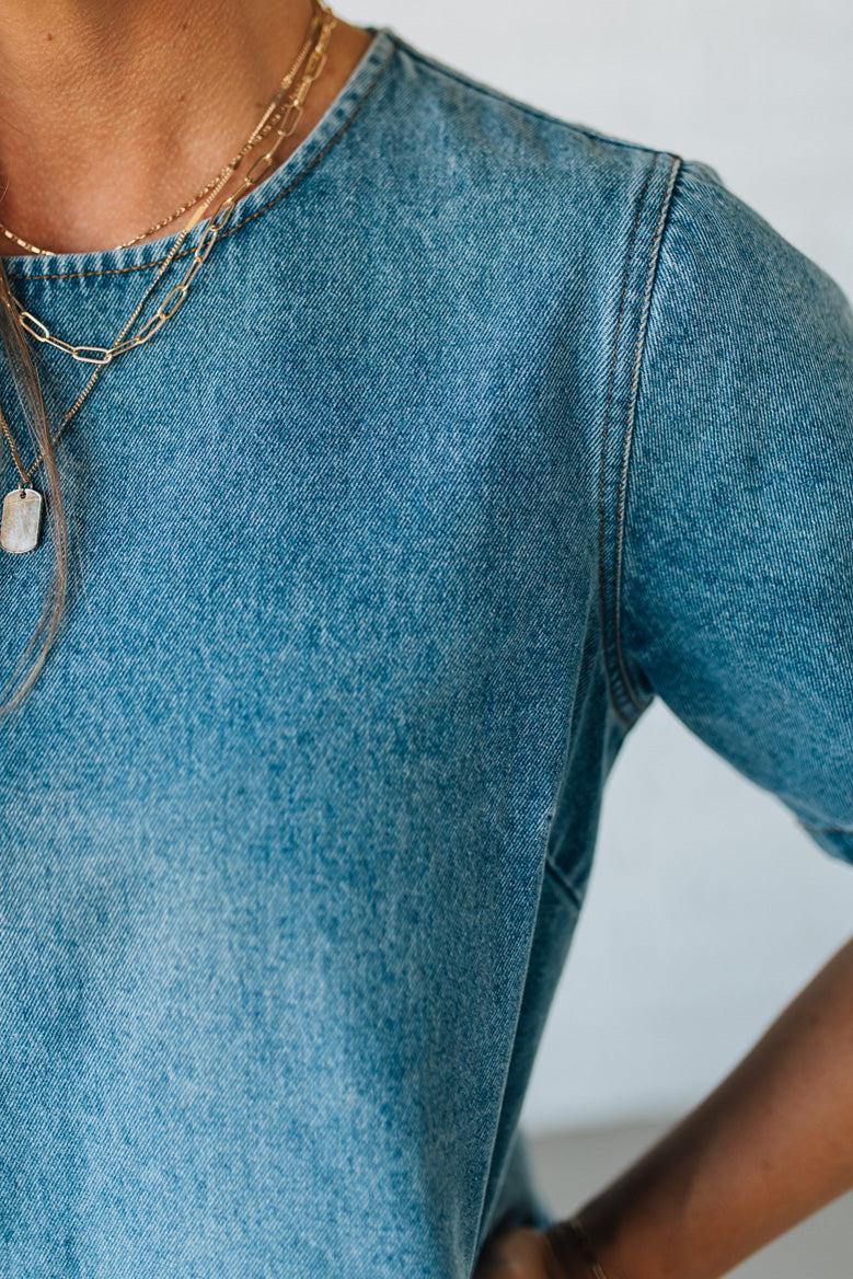 Blonde woman wearing a denim shift dress to the knee with short sleeves, side pickets and bow accents on each pocket.